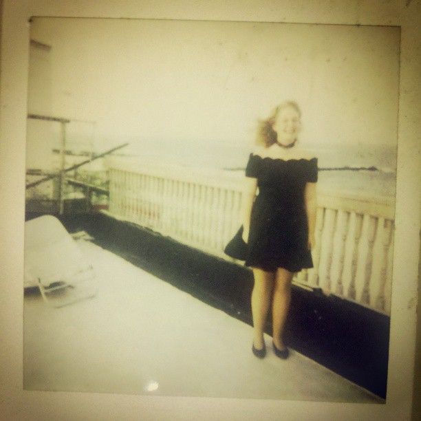 a Polaroid of 13 year old blonde chubby teenager in the 90s wearing a fancy black dress, black flats, and a black choker. She is standing on a disheveled rooftop of building, and the ocean is behind her.